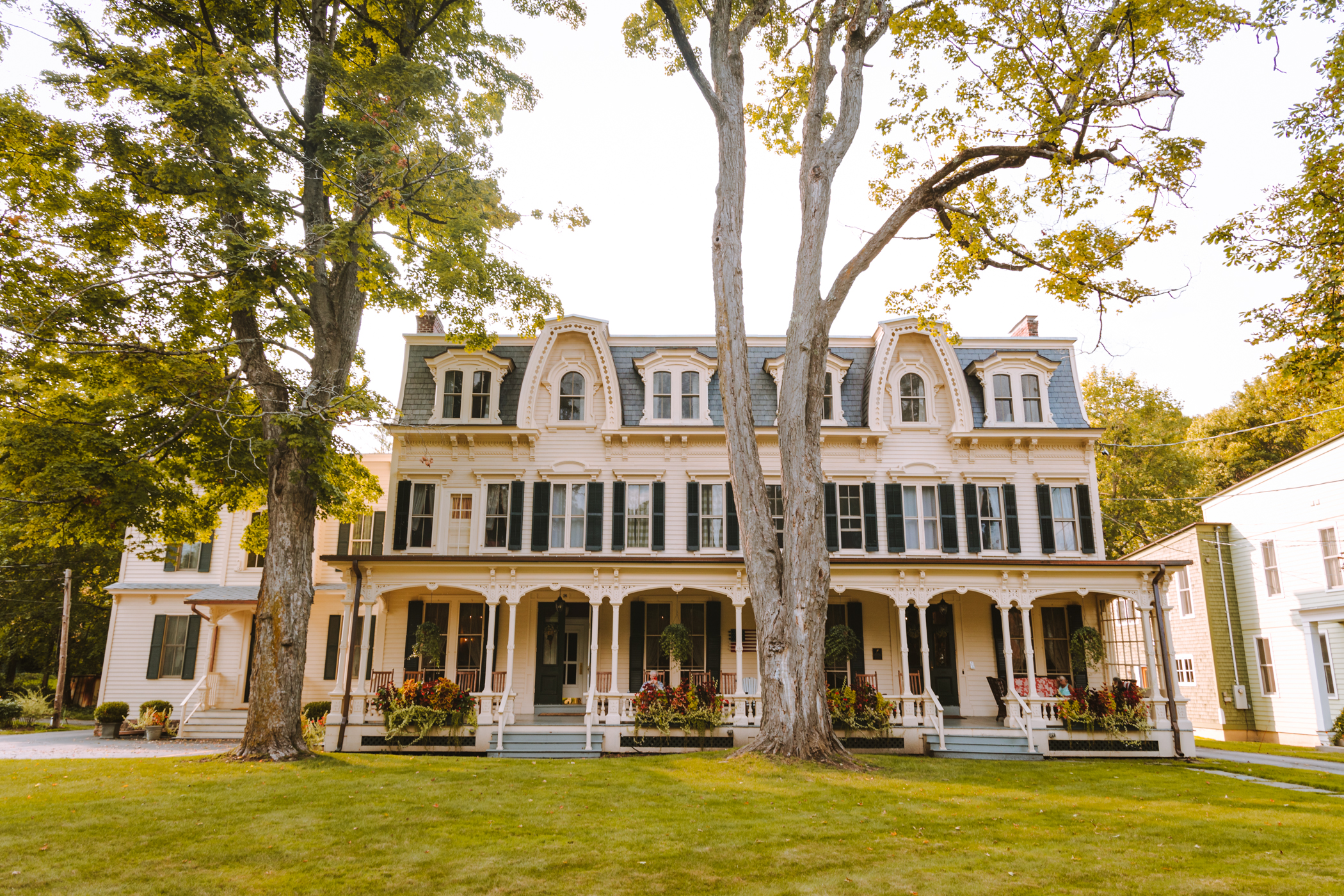 A large, historic Victorian house with ornate details, tall windows, and a wraparound porch, surrounded by green lawn and tall trees in front.